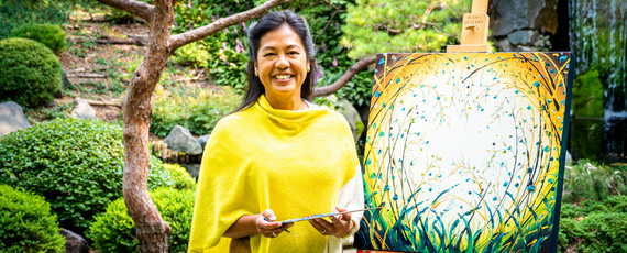 Pamela Sukhum holding a palette in front of her yellow painted canvas in an Arb garden 